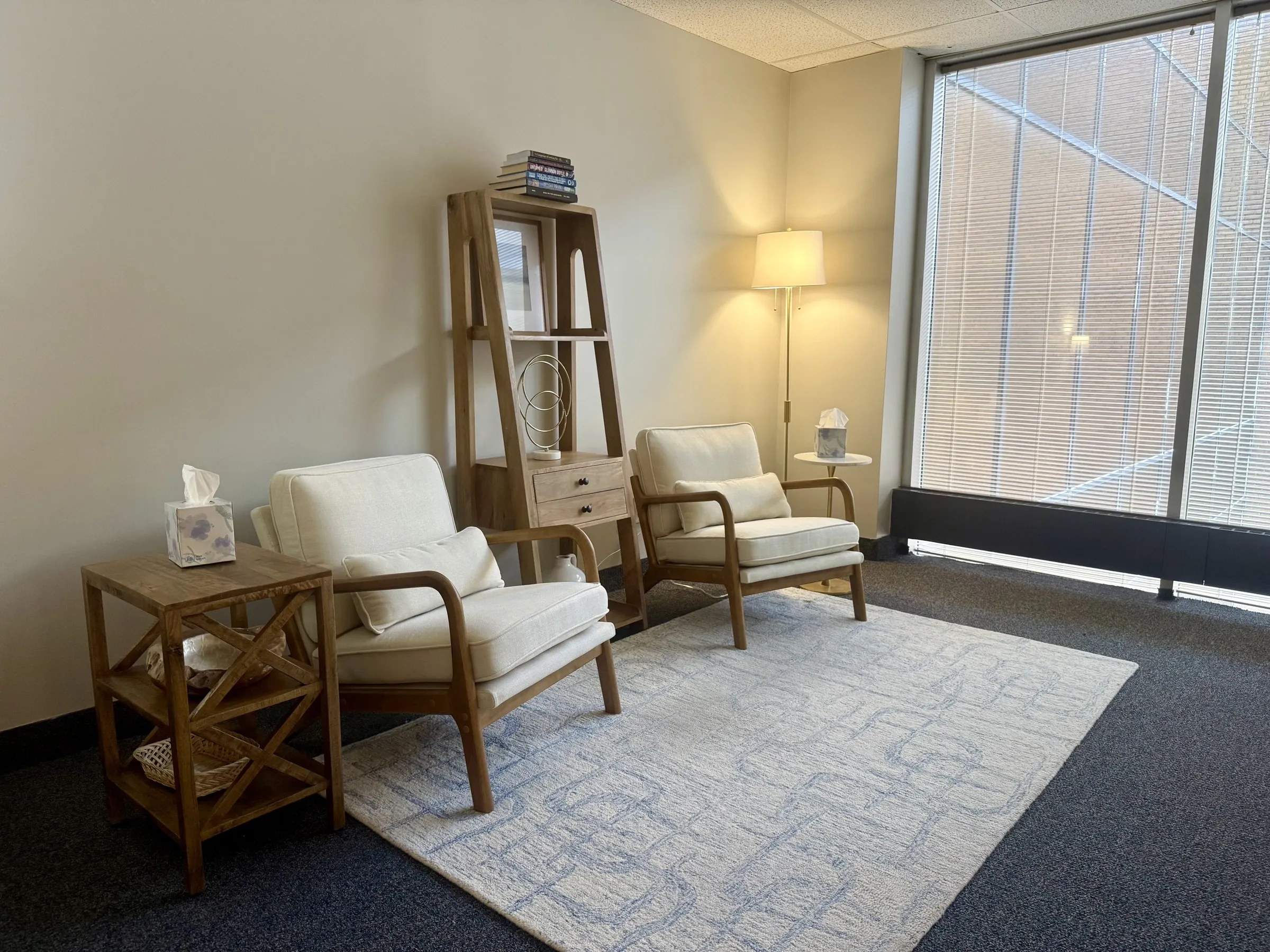 Two cream armchairs on a soft blue rug beside a wooden bookshelf and a large window