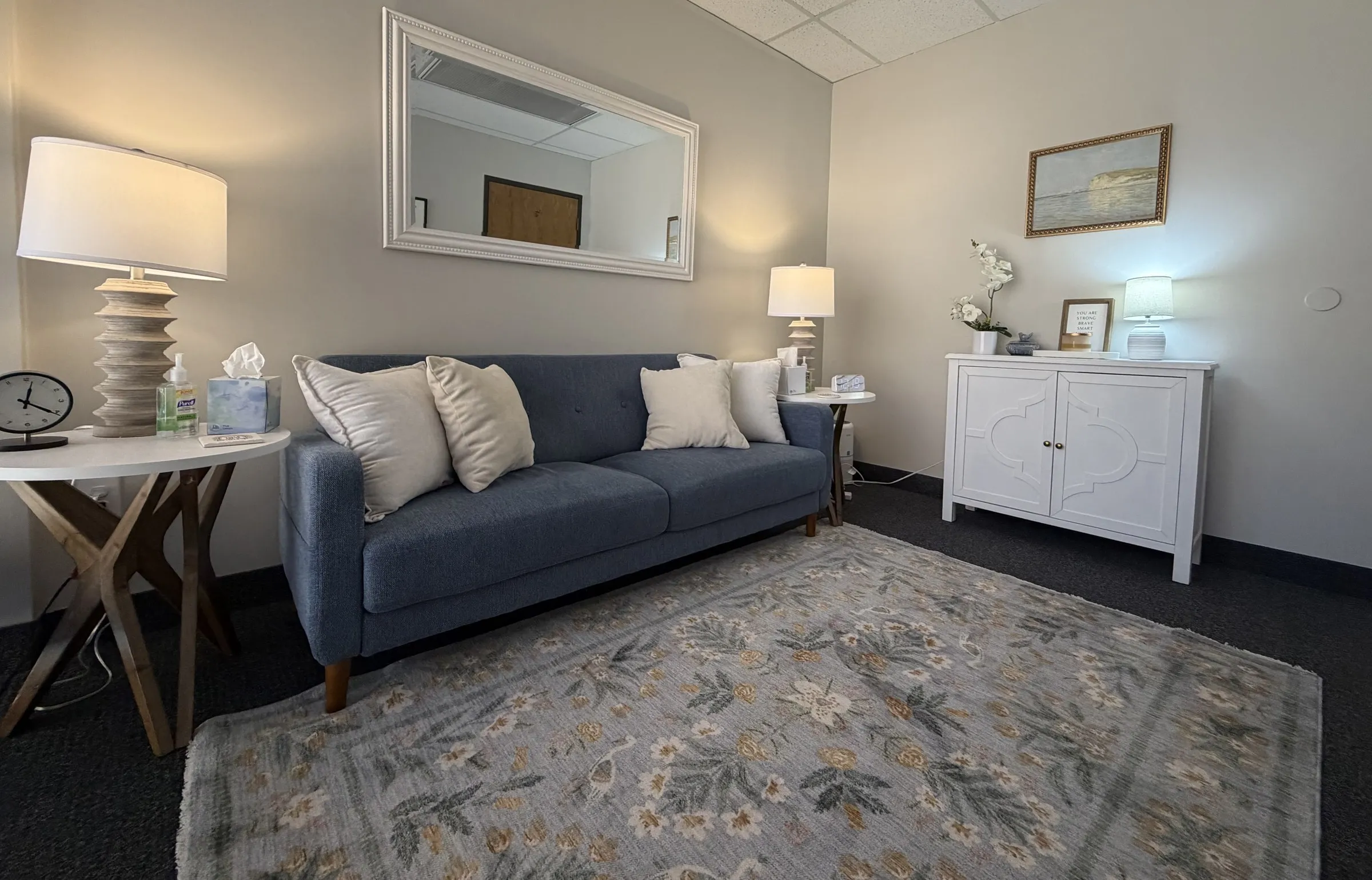 Therapy room with a blue sofa, cream pillows, warm lamps, a floral rug, and a white cabinet